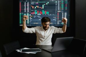 Man in white shirt celebrating a successful trade in front of a large screen displaying stock market charts and data.