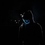 A mysterious hooded figure wearing a glowing neon blue Guy Fawkes mask stands in a dark cyberpunk urban night scene with subtle streetlight glows.