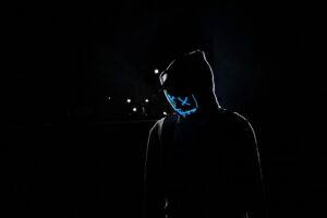 A mysterious hooded figure wearing a glowing neon blue Guy Fawkes mask stands in a dark cyberpunk urban night scene with subtle streetlight glows.