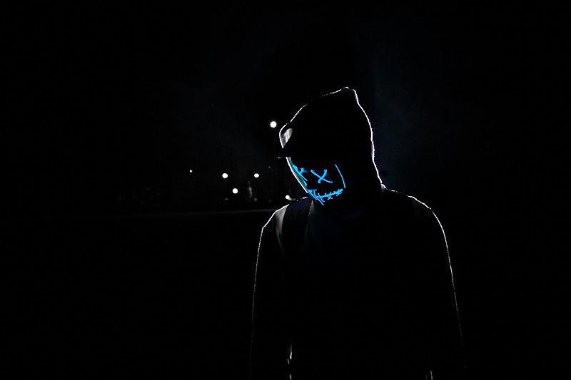 A mysterious hooded figure wearing a glowing neon blue Guy Fawkes mask stands in a dark cyberpunk urban night scene with subtle streetlight glows.